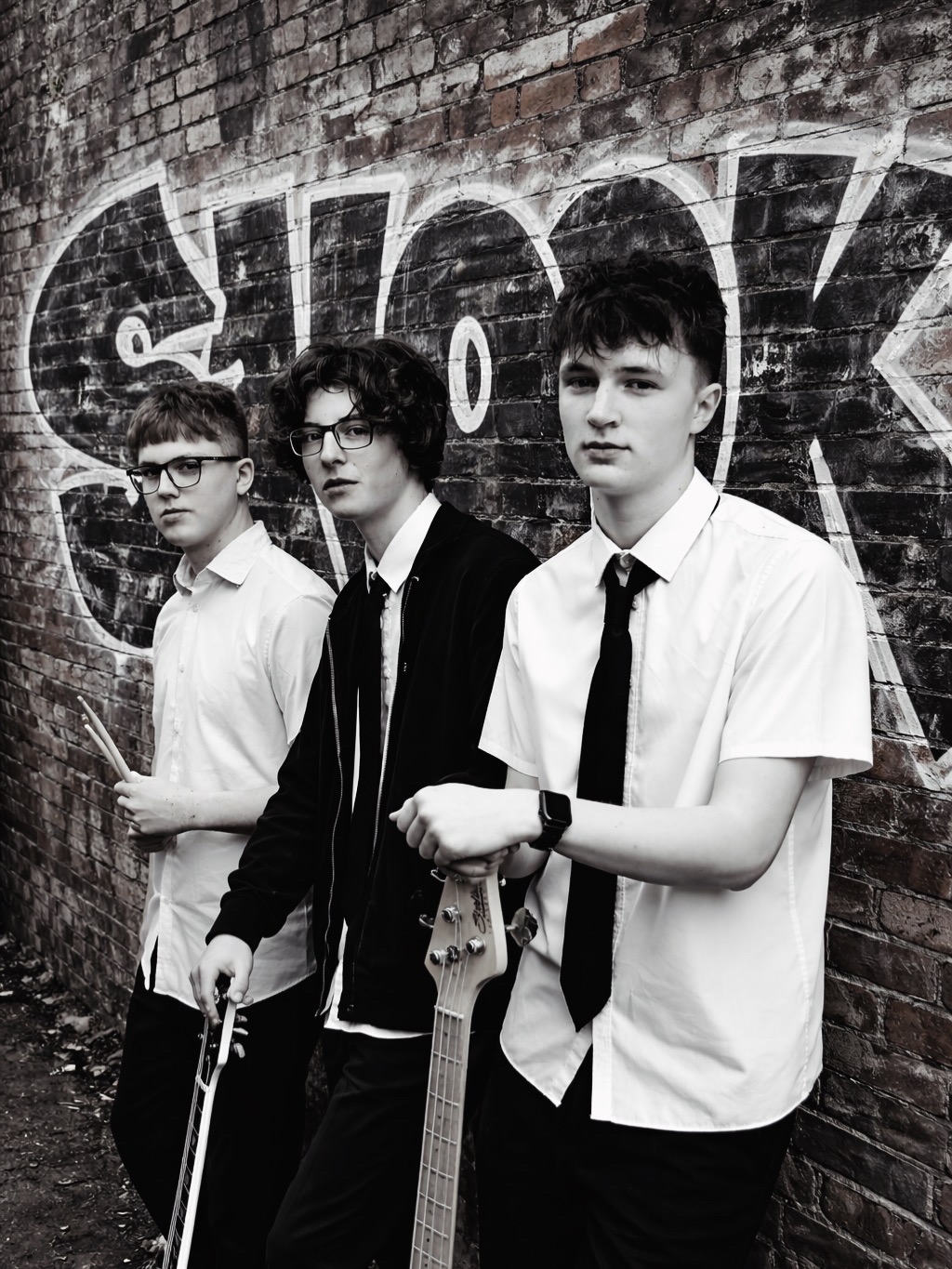 Black-and-white portrait of three band members with instruments in front of a graffiti-covered brick wall.