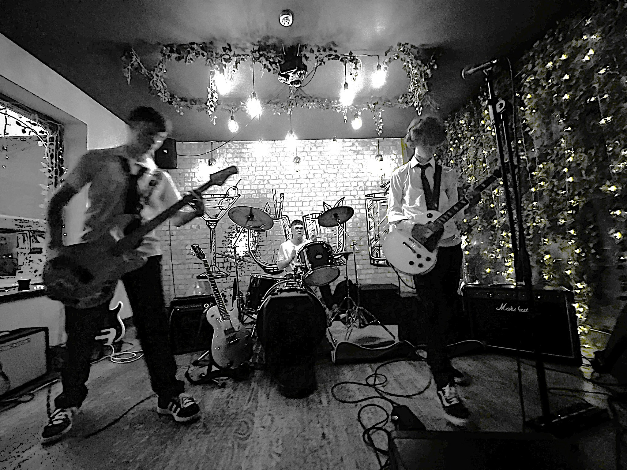 Black-and-white photograph of a three-piece band performing with guitars and drums in an intimate venue.
