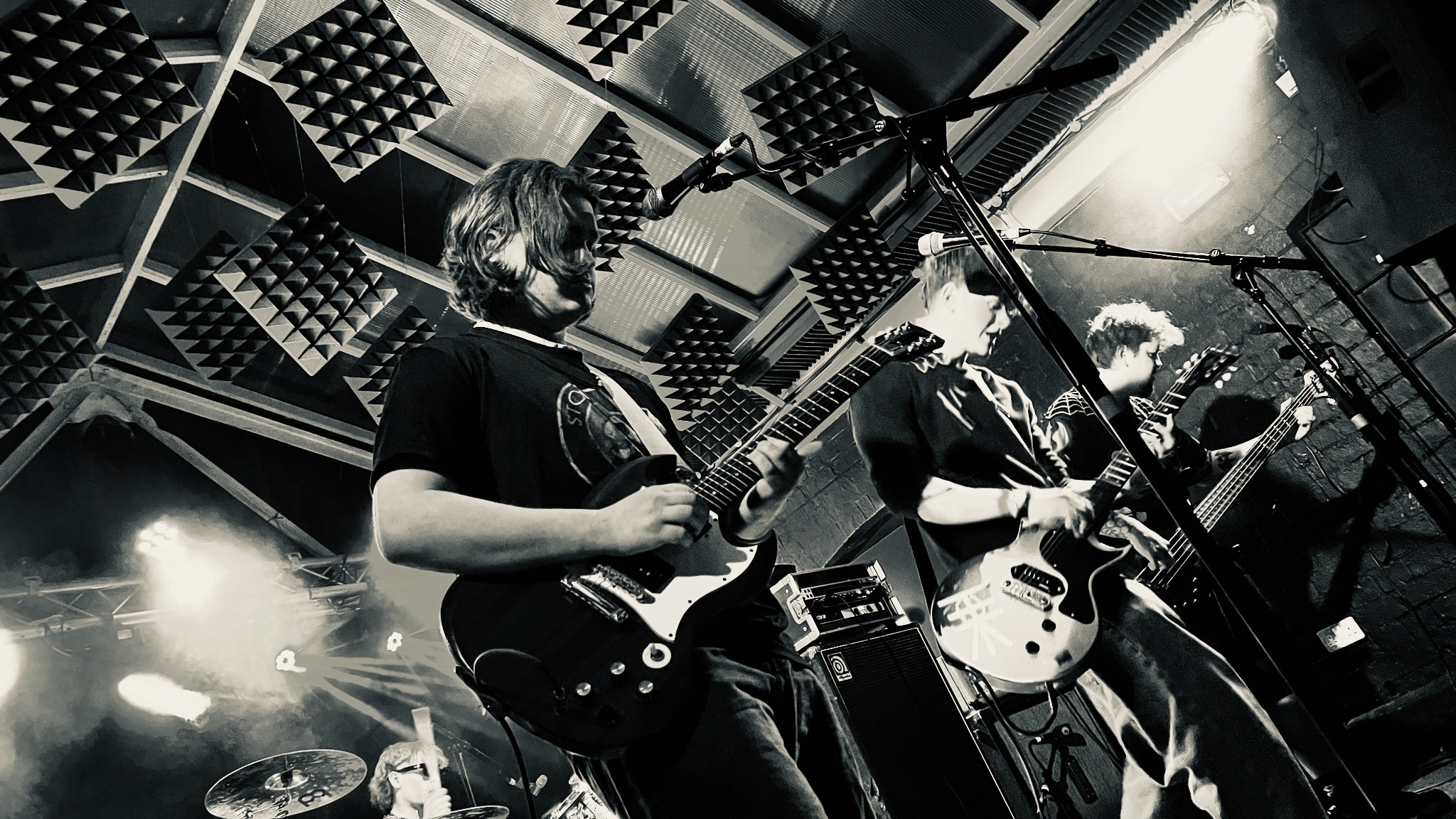 Black-and-white photograph of guitarists and a bassist performing beneath acoustic ceiling panels.